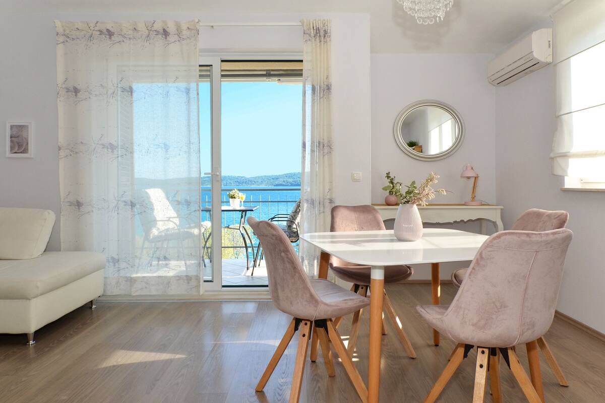 A modern living room with a sofa, a dining table with chairs, a balcony with a view of the sea, and an air conditioning unit.