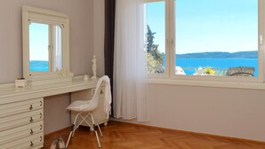 Una habitación con piso de madera, un aparador blanco con cajones, una silla blanca, un ventanal con vista al mar y una cortina blanca.