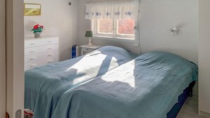 Ein Schlafzimmer mit Bett, Kommode, Nachttisch, einem Fenster mit Vorhang und einer Wandlampe.