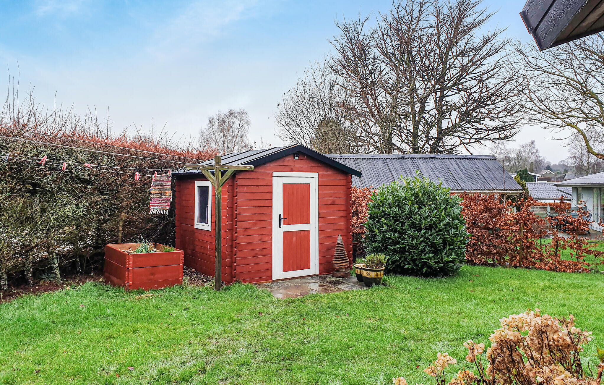 Un petit abri en bois rouge, avec une porte blanche, entouré d’une pelouse bien entretenue et de diverses plantes.