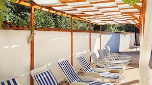 Een rij ligbedden onder een pergola met een betegelde vloer, omringd door een lage muur en groen.