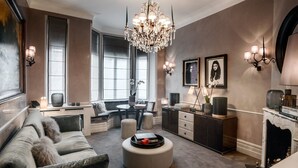 A modern living room with a large chandelier, a sofa, a round ottoman, a fireplace, and a dining area with a table and chairs. - Cadogan Square - 1 bed (London)