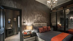 A modern bedroom with a large bed, a nightstand, a mirror, and a chandelier. - Cadogan Square - 1 bed (London)