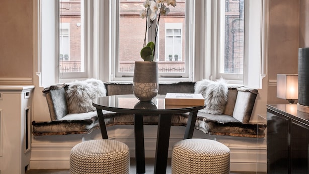 A cozy seating area with a round table, a vase with flowers, and a view of a brick building through large windows. - Cadogan Square - 1 bed (London)