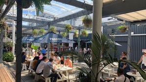 Un café en plein air avec des tables et des chaises, entouré de plantes suspendues et de parasols.