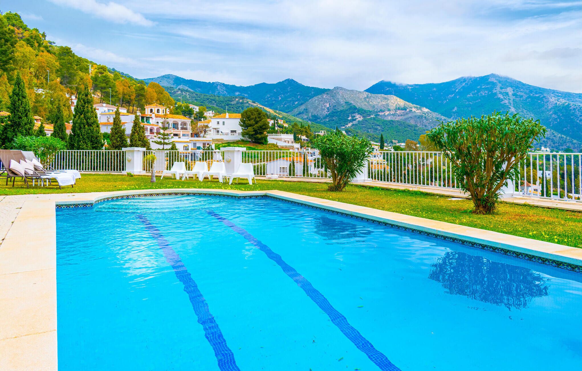 A swimming pool with clear blue water, surrounded by white lounge chairs and a white fence. In the background, there are buildings with red roofs and a mountainous landscape.