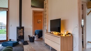 Ett mysigt vardagsrum med en öppen spis, en tv på ett träbord och utsikt över naturen genom ett stort fönster.