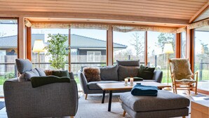 Ett mysigt vardagsrum med en soffa, en pall och en fåtölj, ett soffbord och utsikt över ett hus genom stora fönster.