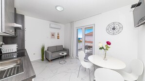 A modern living room with a grey sofa, a small round table with two white chairs, a white air conditioning unit, and a decorative wall clock.