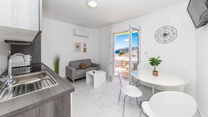 A modern kitchen with a built-in stove, a sink, and a countertop. There is a grey sofa with a brown cushion, a small white table, and a potted plant. A white dining table with chairs is visible, and a large window offers a view of the sea and mountains.