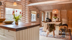 Interior de una cabaña de troncos con una mesa de comedor de madera, un florero con flores rojas, una silla tejida y una alfombra.