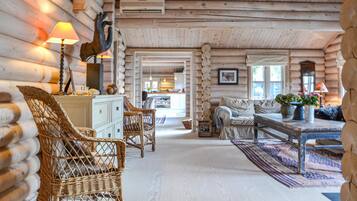 L’intérieur d’une cabane en rondins, avec des meubles en osier, un coin repas et une cuisine visible à travers une porte ouverte.