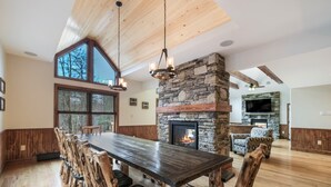 A spacious dining area with a long wooden table and chairs, a stone fireplace, and a large window offering a view of trees. - Rainier by PMR, 8BR, Elevator, Outdoor Fireplace, Double Kitchen, Sleeps 26 (Lake Harmony)