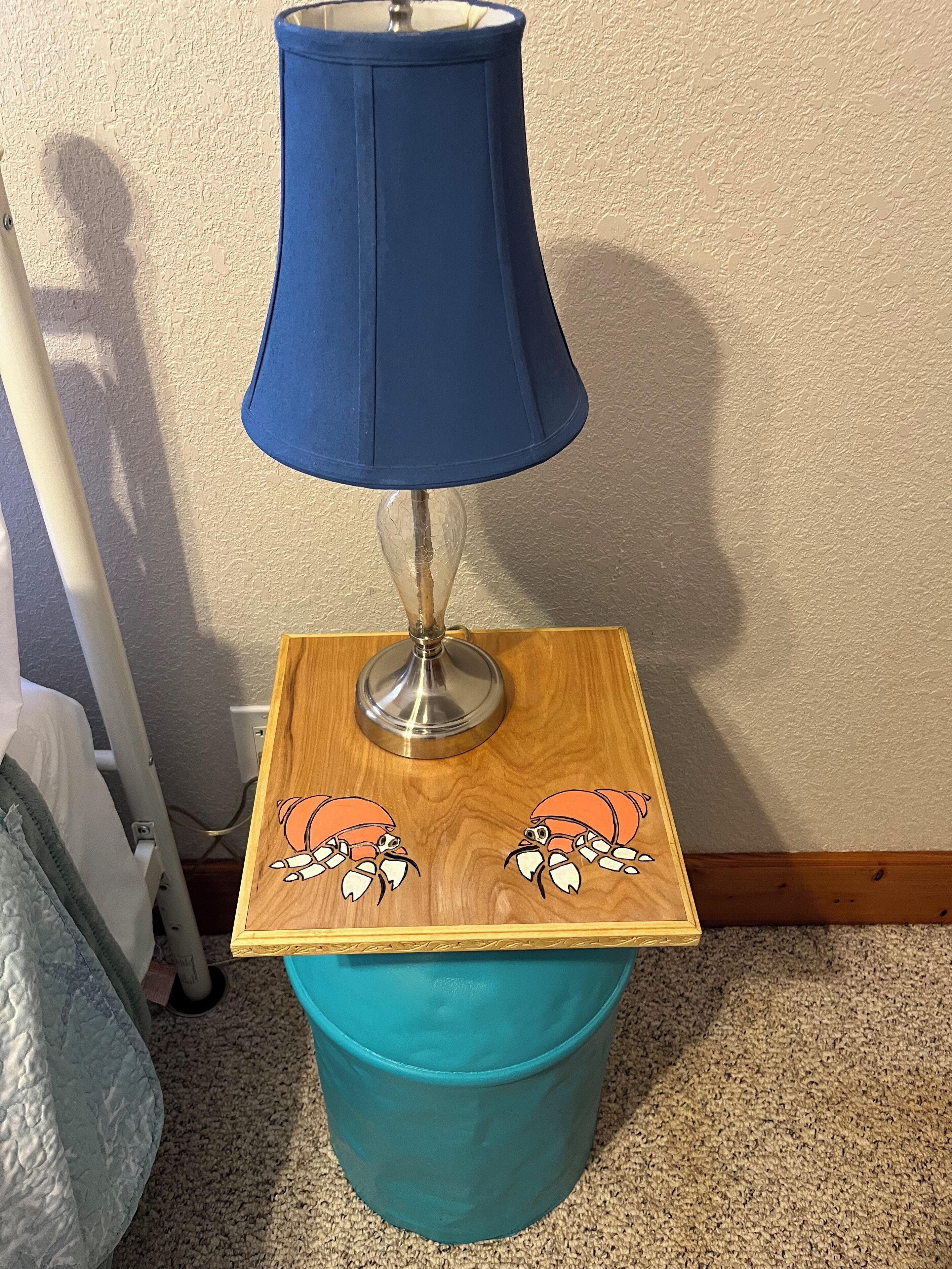 A bedside table with a blue lamp, a wooden side table with crab illustrations, and a teal trash bin.