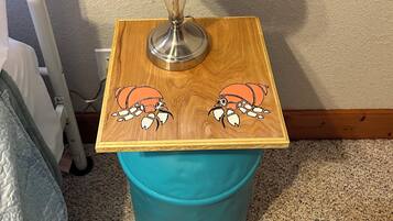 A bedside table with a blue lamp, a wooden side table with crab illustrations, and a teal trash bin.