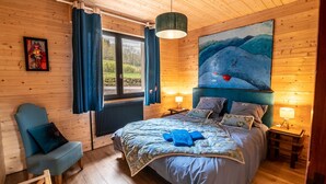Un dormitorio de cabaña de madera con una cama, una silla, un cuadro y una ventana con cortinas.