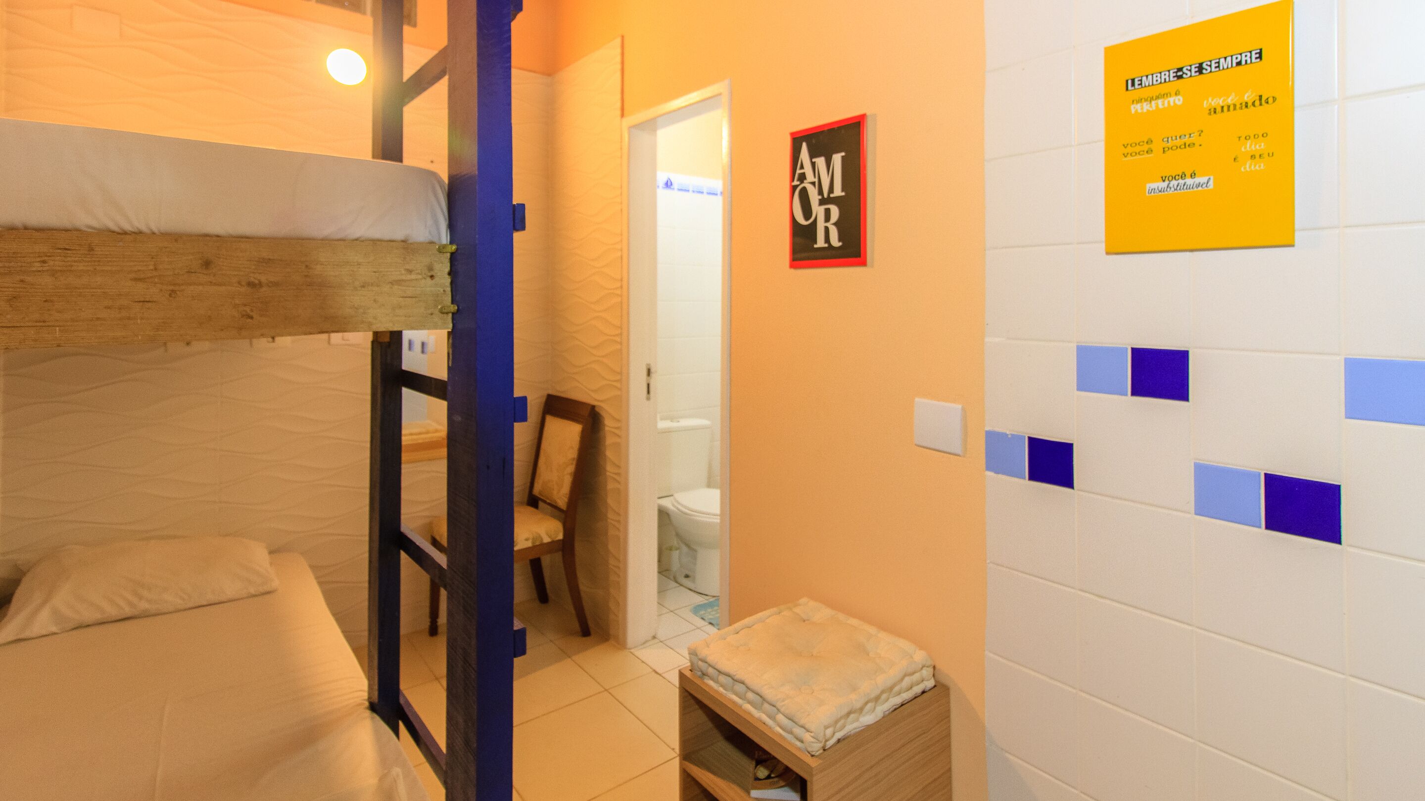 Habitación tipo dormitorio con literas, un taburete de madera, una silla y un baño visible a través de una puerta abierta.