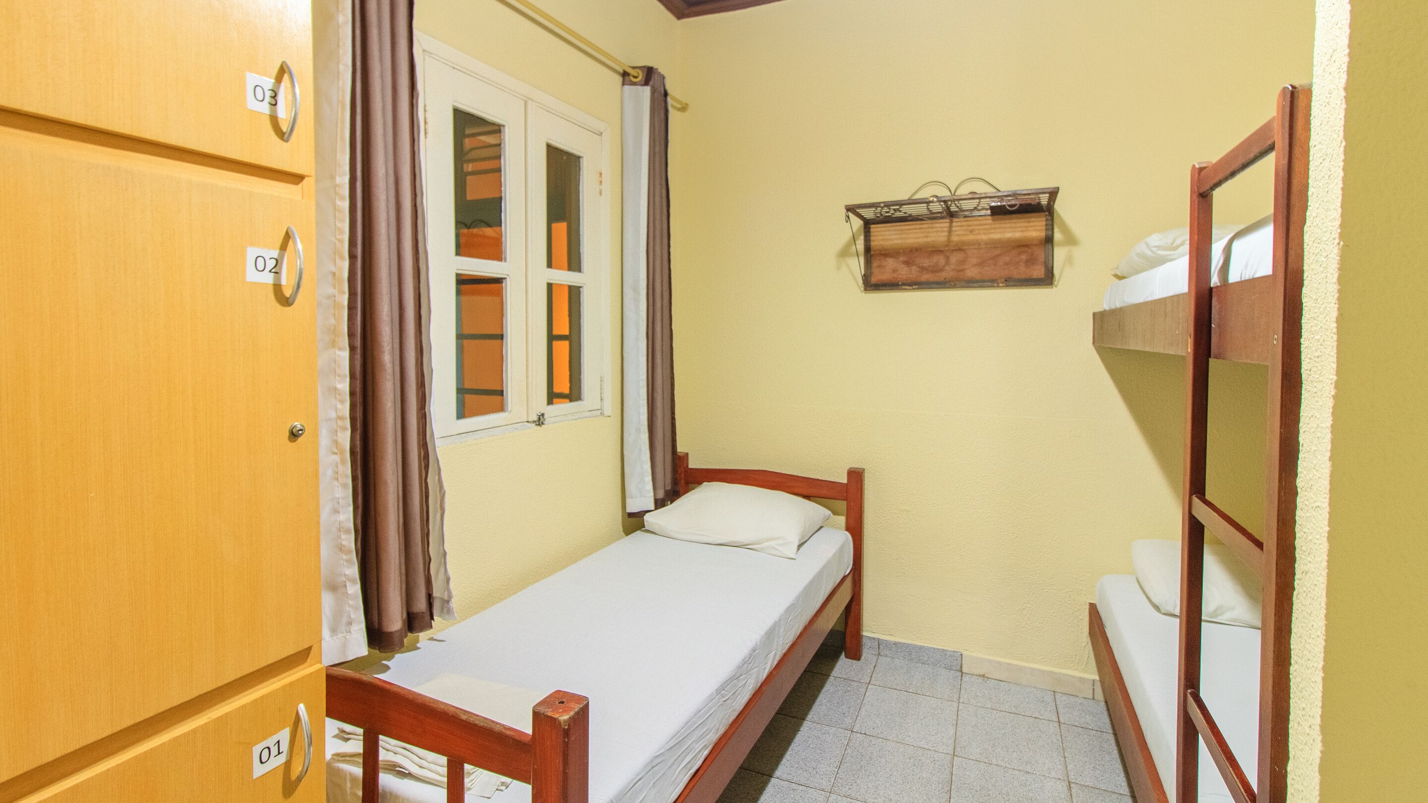 Une petite chambre avec un lit simple, une armoire en bois, une fenêtre avec des rideaux, une table de chevet et une étagère fixée au mur.
