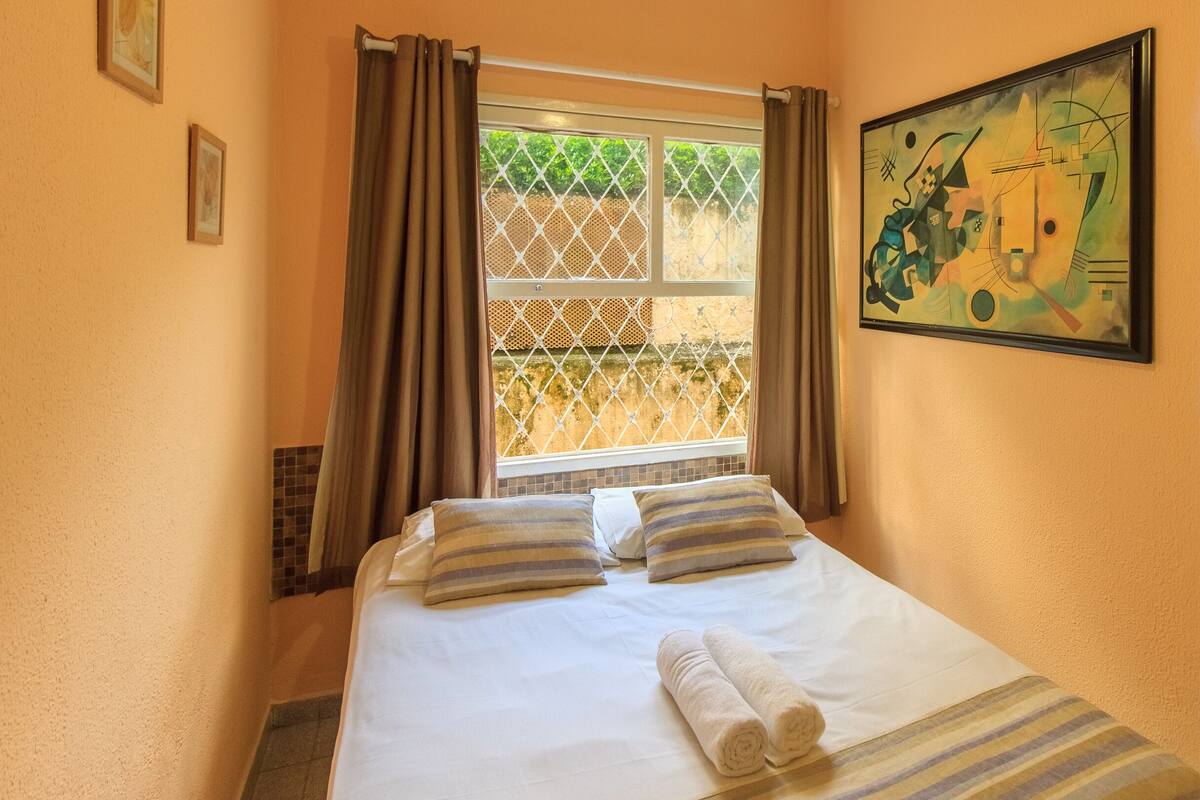 Une chambre d’hôtel avec un lit, deux oreillers, une fenêtre avec des rideaux et un tableau au mur.