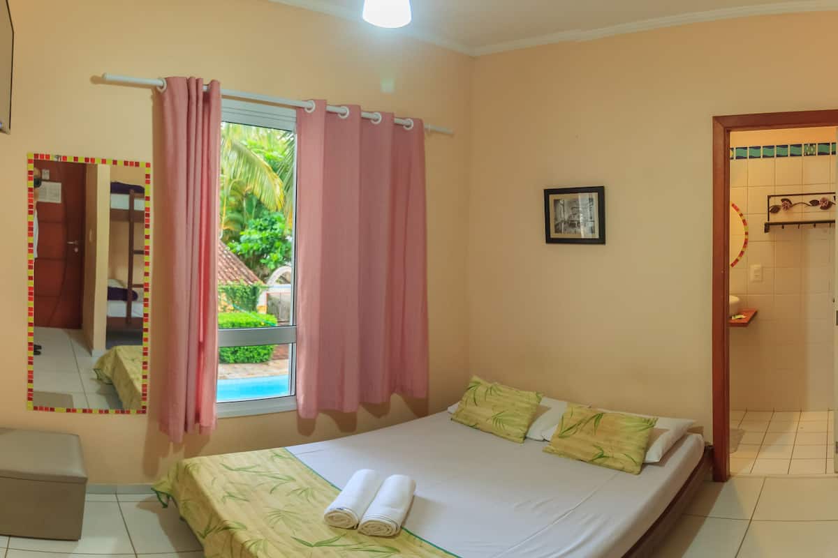 Une chambre à coucher comprenant un lit, une fenêtre avec des rideaux, une table de chevet, un miroir et une porte donnant sur une salle de bain.