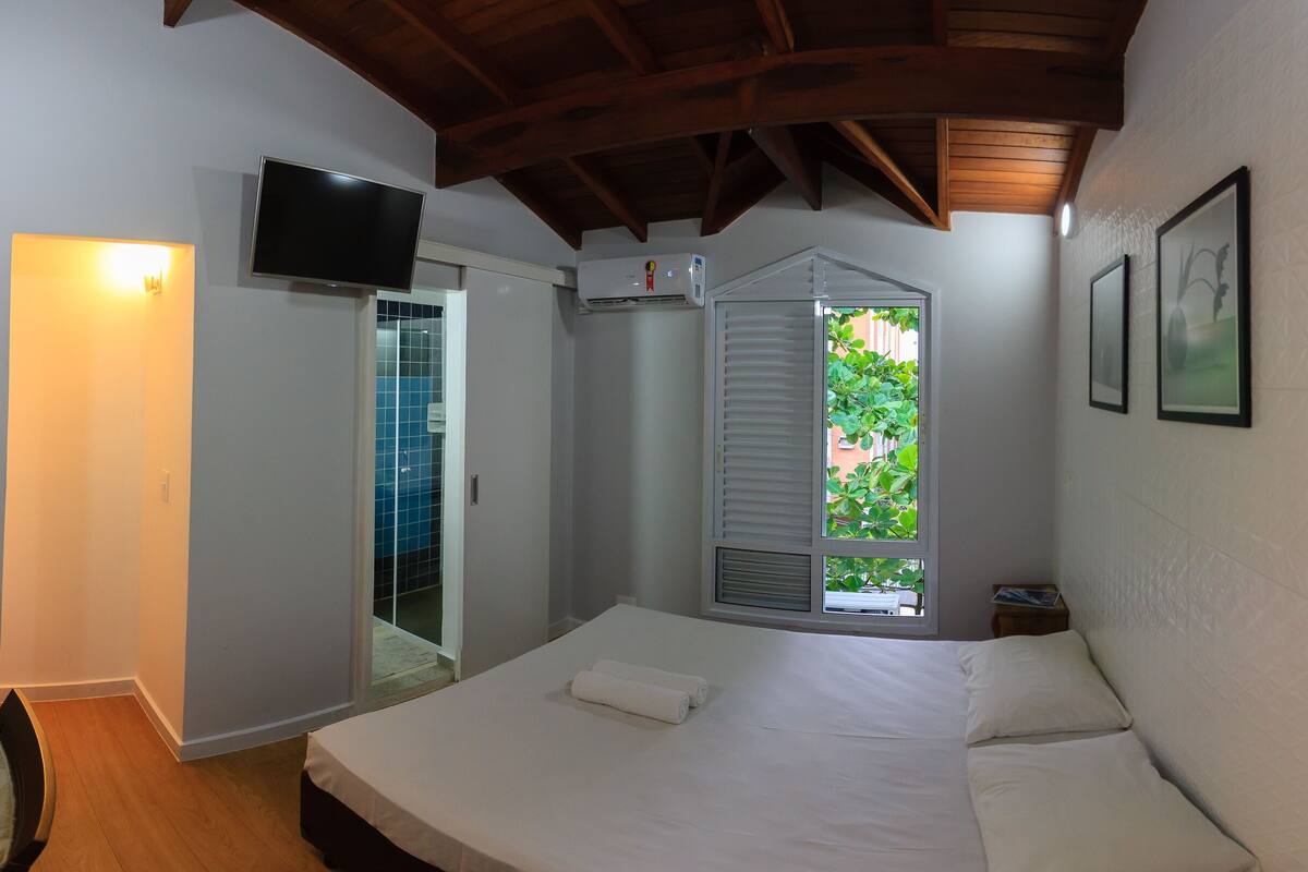 Une chambre à coucher comprenant un lit, un téléviseur, une fenêtre avec des stores et une porte donnant sur une salle de bain.