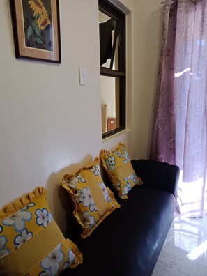 A living room with a dark sofa, two yellow pillows with blue floral patterns, a framed picture on the wall, a window with a view, and a curtain with a floral pattern.