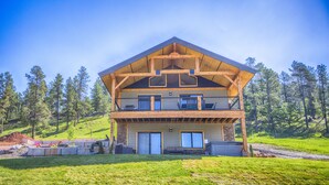 A two-story wooden house with a balcony, surrounded by a grassy area and trees. - Crystal clear Mountain View’s with a farmhouse chic vibe. (Sturgis)