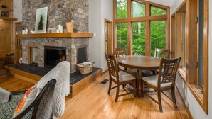 Una sala de estar acogedora con chimenea de piedra, mesa de comedor de madera y ventanas amplias con vistas a un bosque.