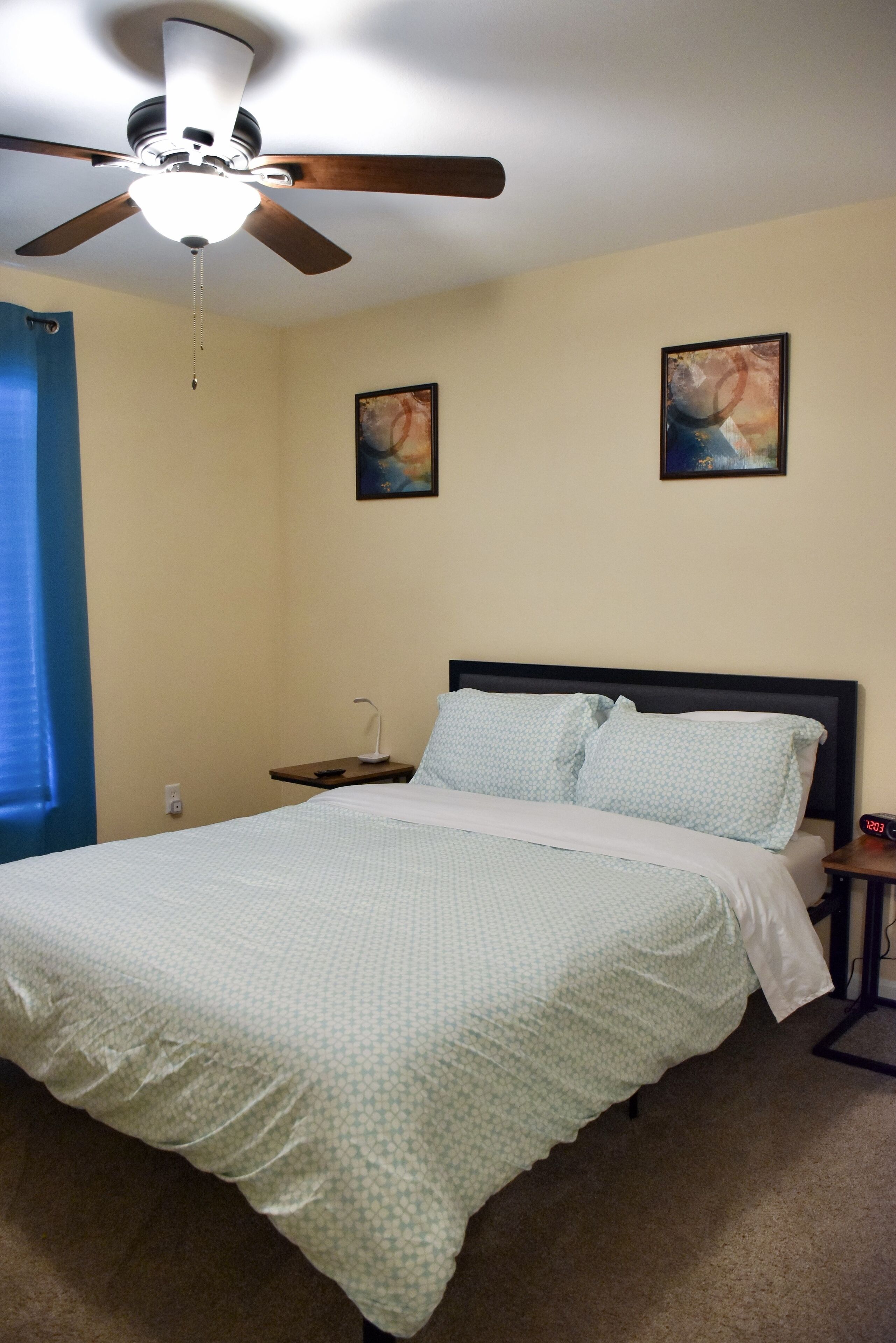 A bedroom with a bed, a ceiling fan, a nightstand, two wall art pieces, and a window with blue curtains.