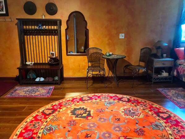 A room with a wooden floor, a round orange carpet, a small round table with a chair, a wooden bench with shoes, a mirror, and a small cabinet.