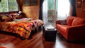 A cozy room with a bed, a sofa, and a small table. There are windows with curtains, a view of greenery outside, and a wooden floor.