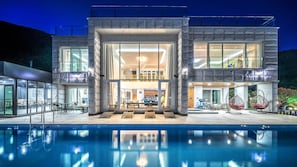 Una casa moderna de dos pisos con un amplio ventanal, una piscina al frente y mobiliario de exterior.
