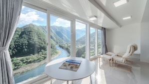 A room with a large window overlooking a river and mountains, a round table with a magazine, and two chairs. - Miramonti Pool Villa Pension (Gapyeong)