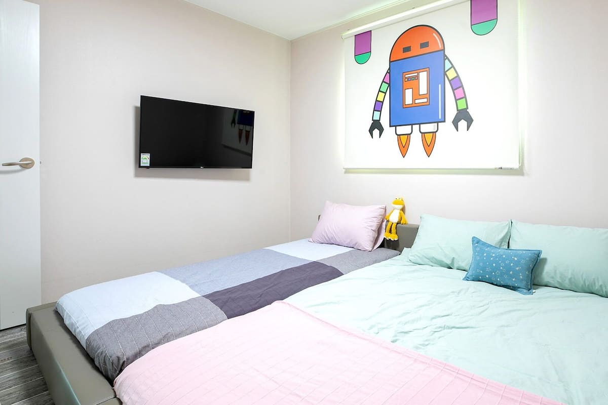 Een slaapkamer met een bed, kussens, een televisie en een wanddecoratie van een cartoonrobot.