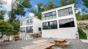 Un bâtiment moderne à deux étages, doté de grandes fenêtres, d’une terrasse en gravier avec des tables de pique-nique et d’un escalier extérieur menant à l’entrée.