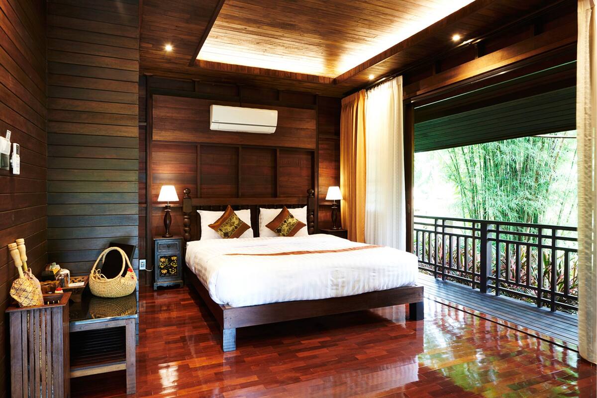 Un dormitorio con paneles de madera, una cama grande, lámparas de noche, una mesita de noche y una ventana con vistas a la vegetación.