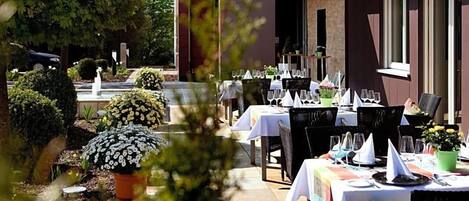 An outdoor dining area with tables set for guests, surrounded by potted plants and a fountain.