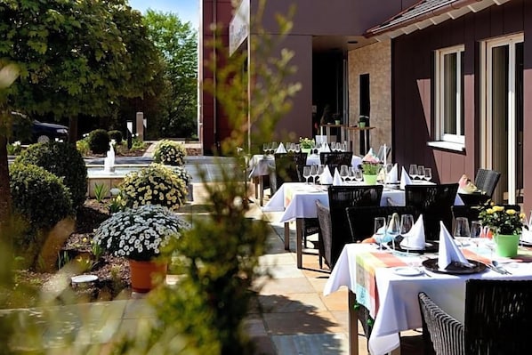 An outdoor dining area with tables set for guests, surrounded by potted plants and a fountain.