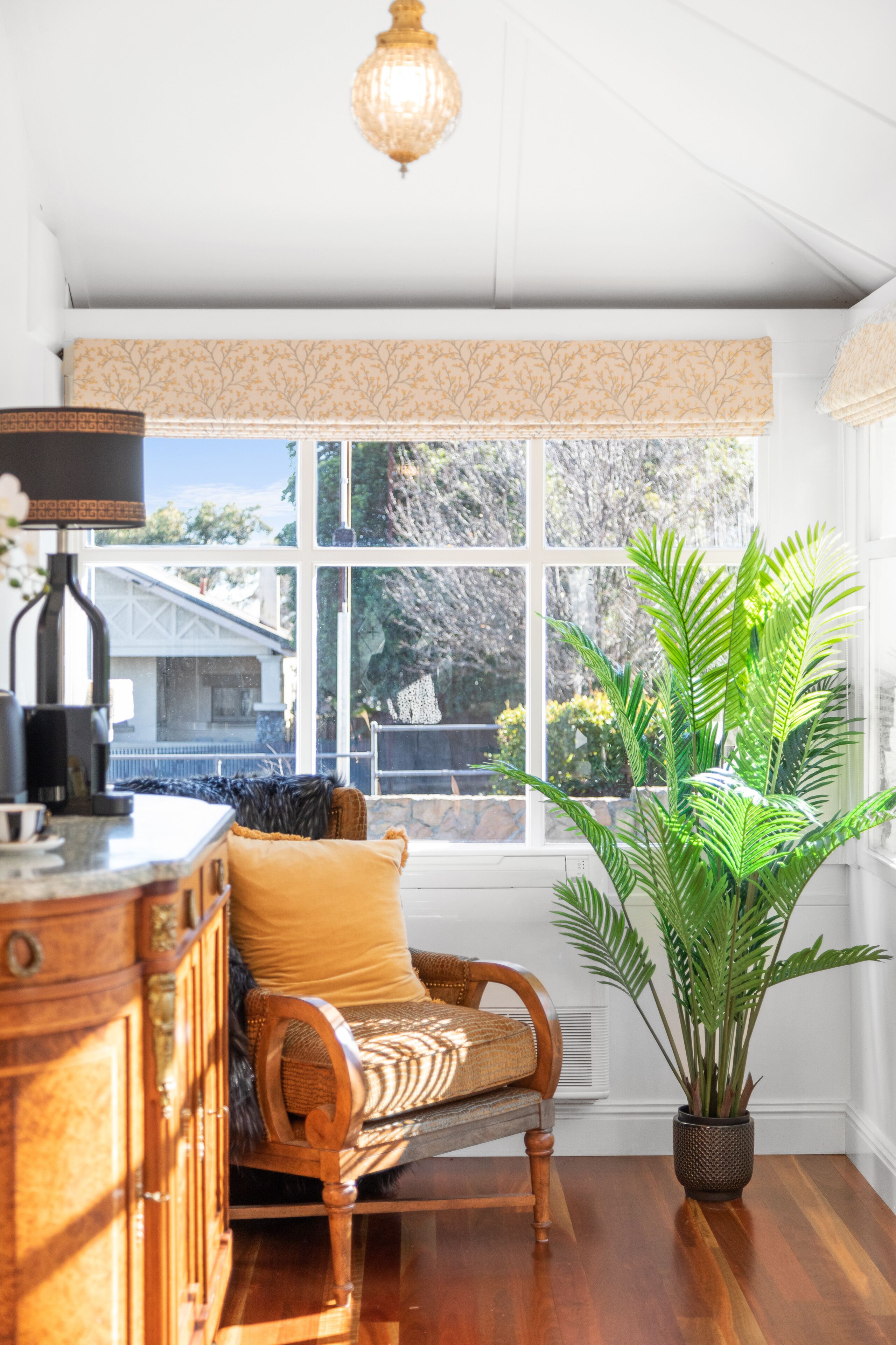 A room with a large window, a wooden chair with a yellow cushion, a potted plant, and a wooden cabinet.