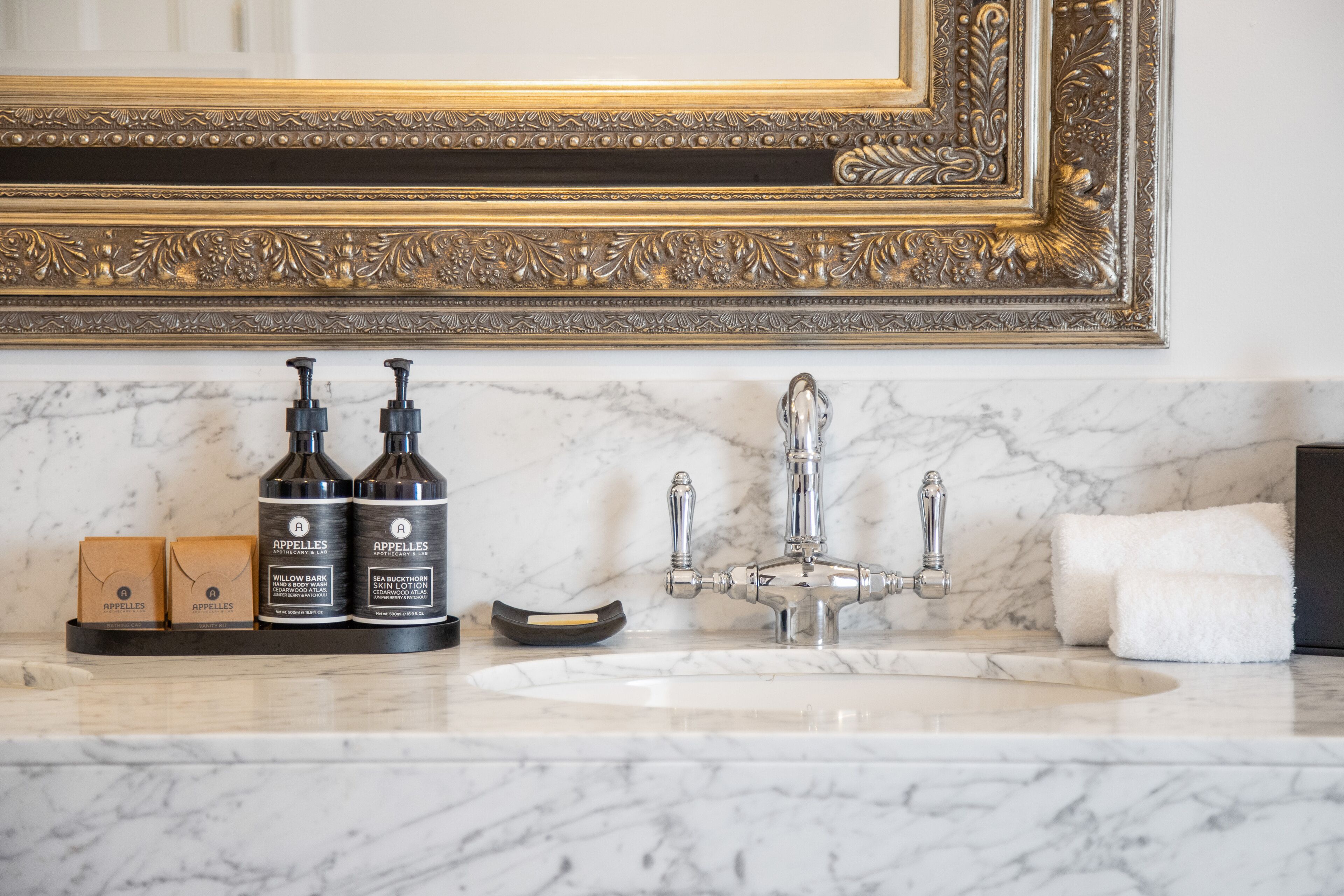 Um lavatório de banheiro com bancada de mármore, um espelho com moldura dourada e duas embalagens de Apelles body wash.