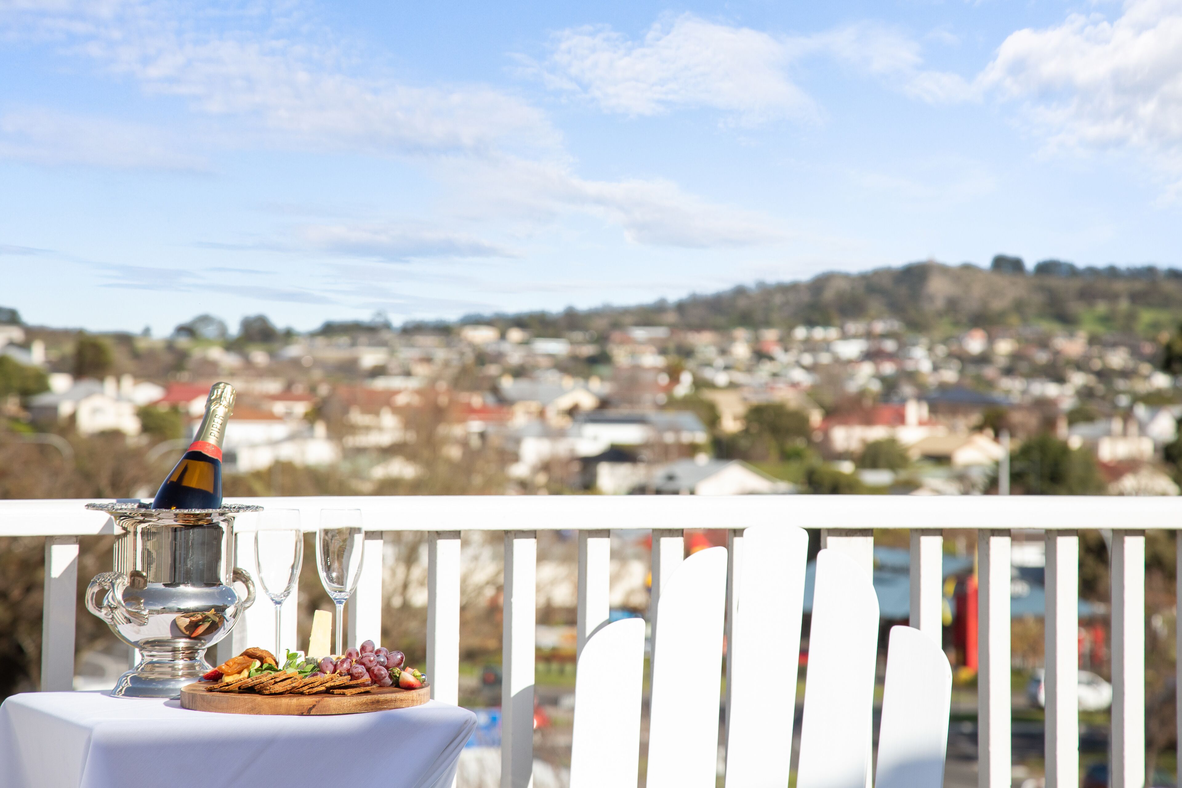 A table with a silver charger set, a bottle of champagne, and a plate of grapes and crackers on a balcony overlooking a residential area.