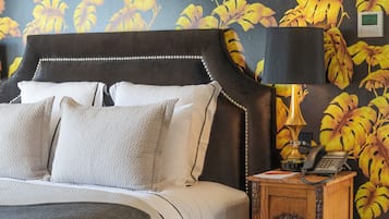 Una cama bien hecha con un cabecero oscuro, una lámpara de noche y un teléfono sobre una mesita de madera.