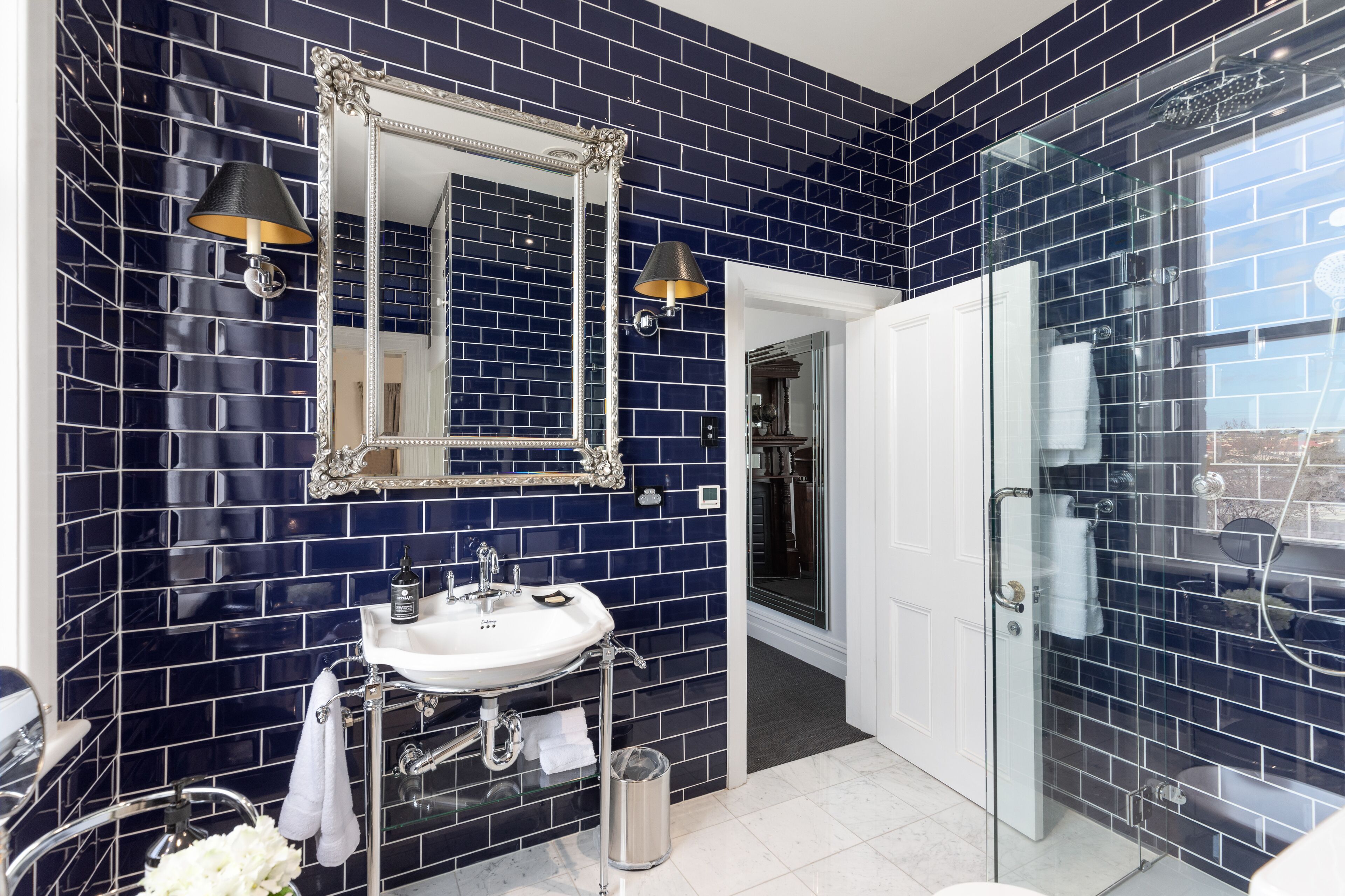 A bathroom with dark blue tiled walls, a white sink, a large mirror, and a glass shower enclosure.