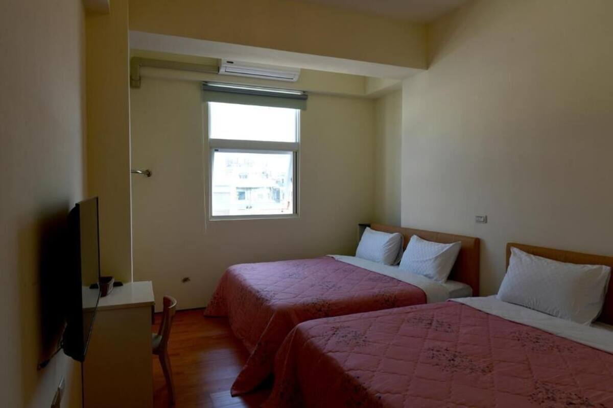 A small, simple room with two beds, a desk, a chair, and a television.