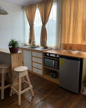 Eine kompakte Küche mit Spüle, zwei Barhocker, einem kleinen Kühlschrank und einem Fenster mit Vorhängen.