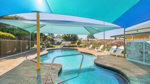 Una piscina con sombrilla, sillas de descanso y un área delimitada por una valla.