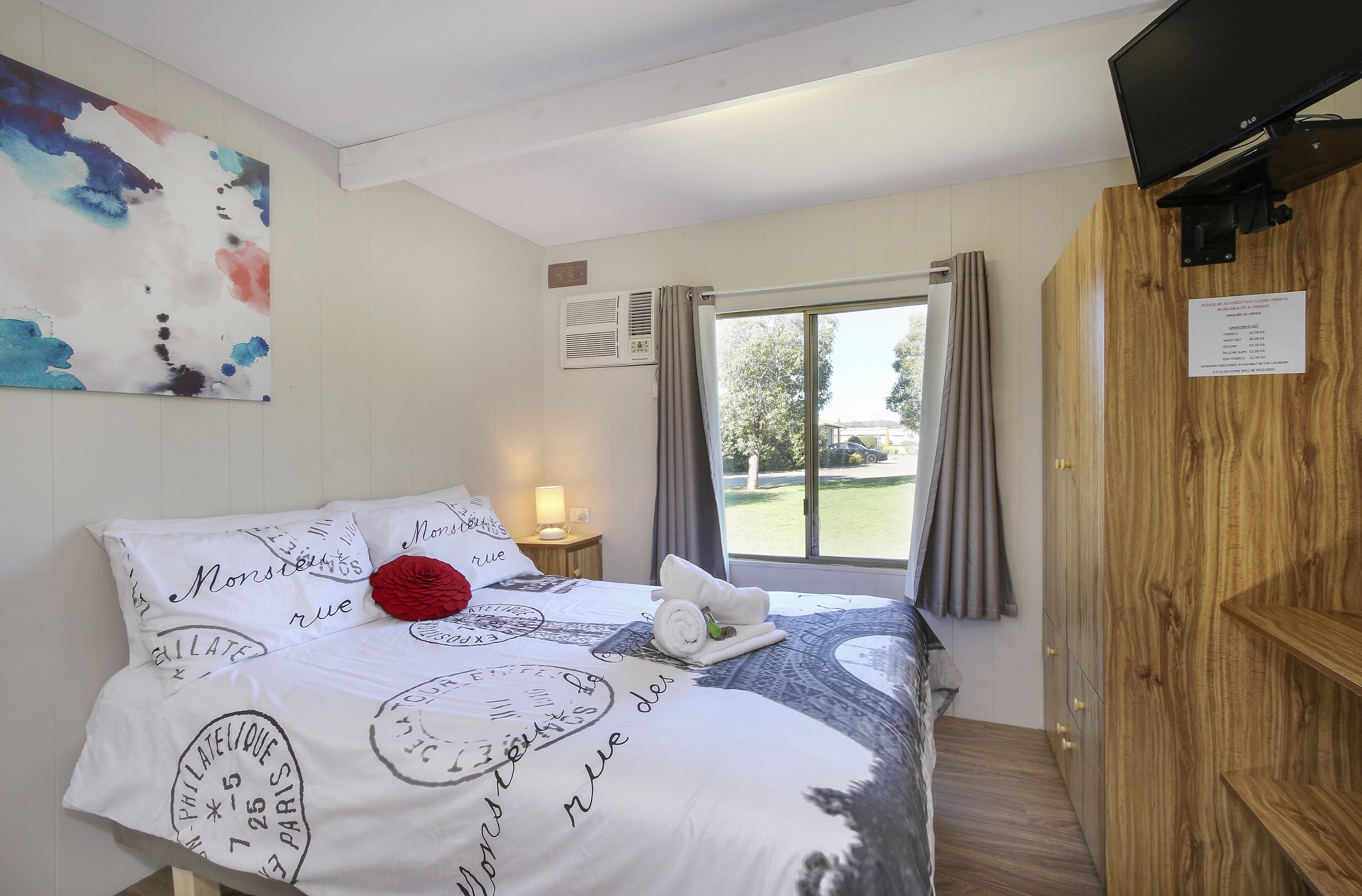 Una habitación de hotel con una cama, una ventana con cortinas, un armario de madera y un televisor fijado a la pared.