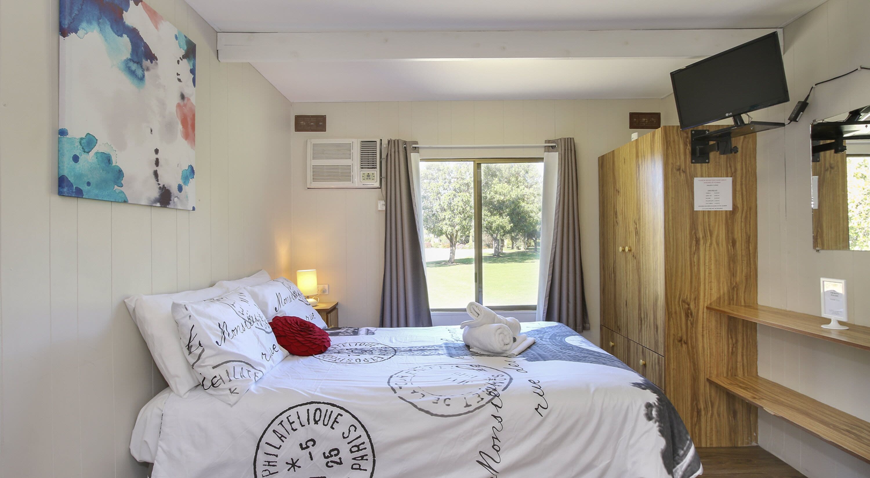 A hotel room with a bed, a television, a window with curtains, a wall-mounted air conditioner, and a wooden wardrobe.