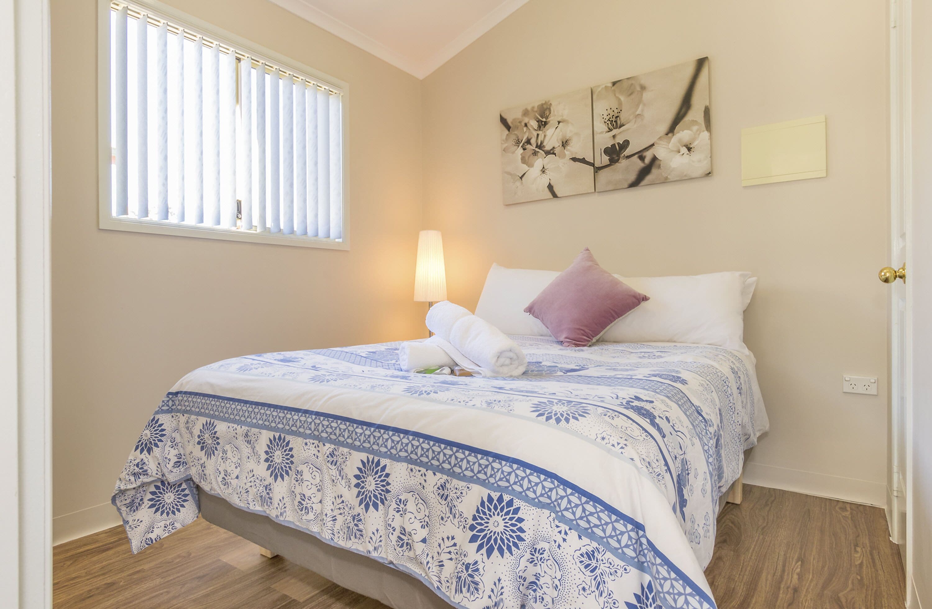Een net opgemaakt bed met een blauw-witte bloemenovertrek, één paars kussen en een witte muur met een bloemschilderij erboven.