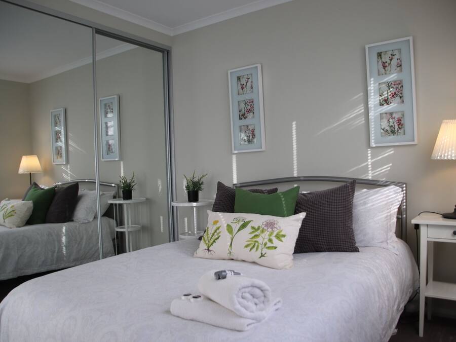 Een net opgemaakt bed met een opgerolde witte handdoek, een bedlampje en een klein tafeltje met een plantje.
